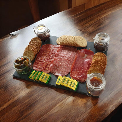 A charcuterie board arranged on a dark slate platter on a wooden table, featuring sliced salami and prosciutto, crackers, olives, small yellow packets, and two glass jars with preserves.