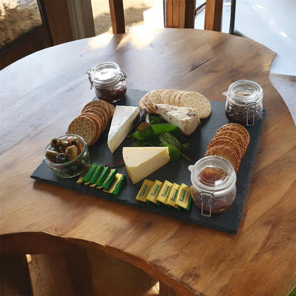 A charcuterie board styled with cheeses, crackers, olives, and jars of preserves on a dark slate serving platter, set on a rustic wooden table.